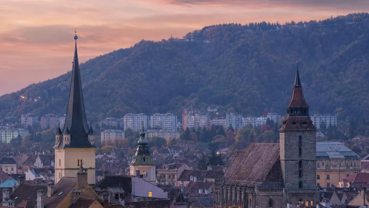 Church spires and tiled roofs in a historic centre at dusk, with apartment blocks at the foot of a wooded hill in autumn colours