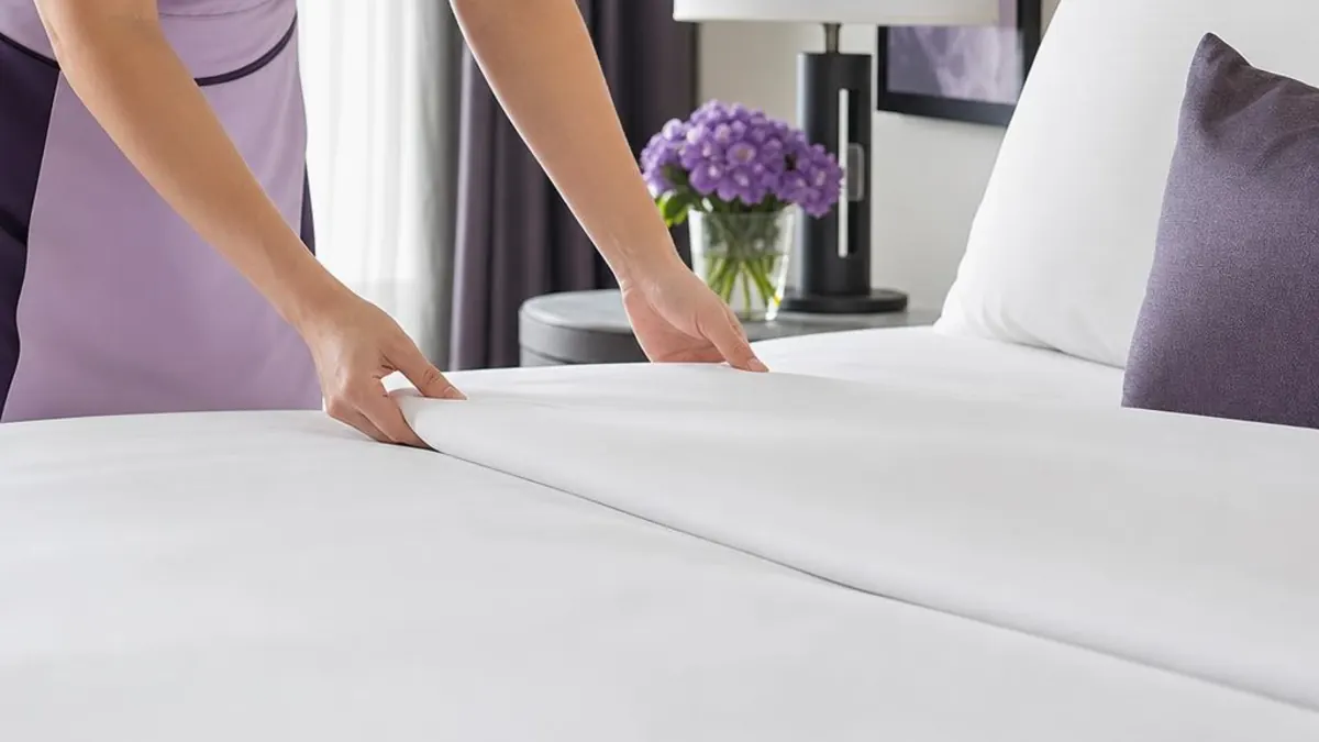 Housekeeper in a lilac uniform smoothing white linen on a bed, next to a bedside table with purple flowers
